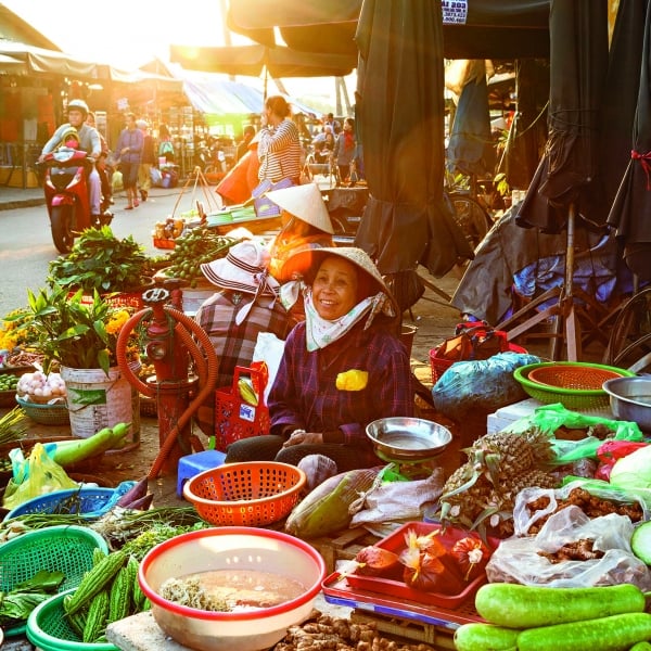 A smiling lady wearing a boater hat sits amongst her wares at a busy outdoor marketplace.