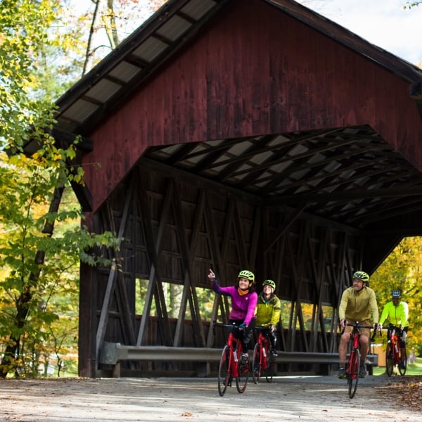 Cyclists biking through a covered bridge, surrounded by fall foliage, on a Vermont bike tour, In Stowe, Vermont